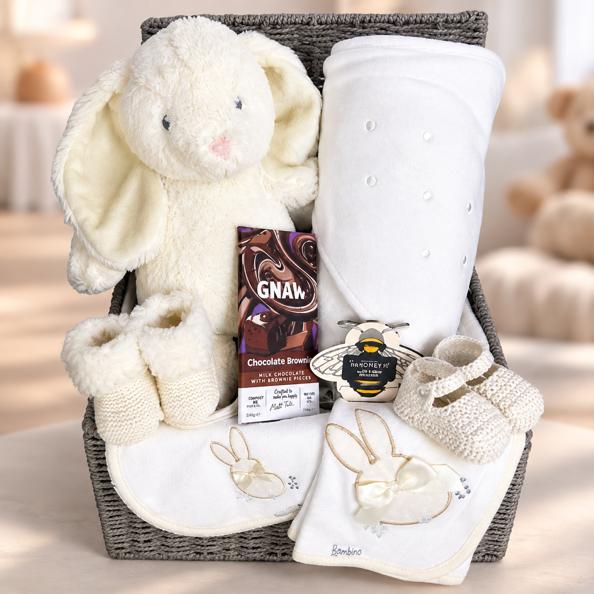 A baby gift basket set containing a plush cream bunny toy, a rolled white hooded baby towel, a pair of cream knit booties, a white bib  and dribble cloth with embroidered bunny details, a pair of baby shoes and a pair of baby booties and a bar of Gnaw chocolate