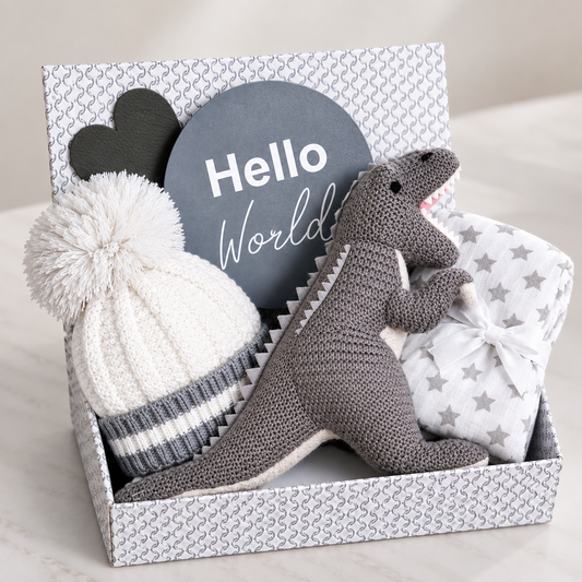 A baby gift box filled with soft items: a grey knitted dinosaur toy, a white knit pom-pom hat with grey stripes, a white baby muslin with grey stars, and a round grey sign that reads 'Hello World' on a light neutral background.