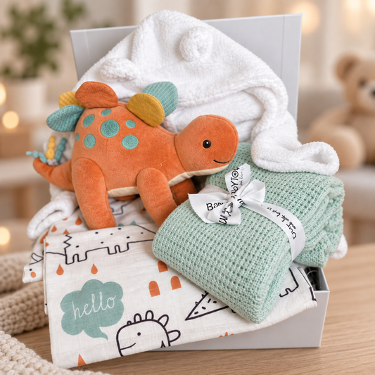 A baby gift hamper in a white baby keepsake box featuring a plush orange dinosaur toy form Mary Meyer, a white baby dressing gown, folded mint green cotton cellular baby blanket, and a patterned baby muslin with dinosaur illustrations.