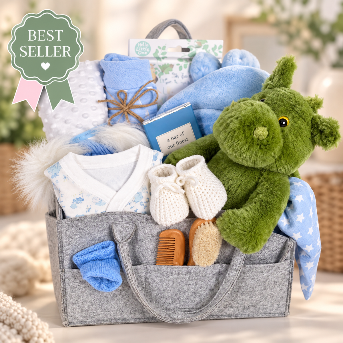 A grey nappy caddy containing a green dragon soft baby toy, a blue and white baby sleepsuit, a blue and white baby pompom hat, a soft white baby blankets, a blue baby dressing gown, a Ziggle baby knot hat and  in blue and white stars.