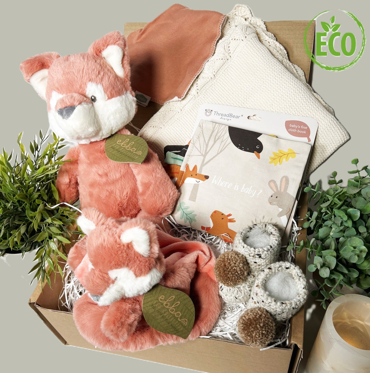 Neutral New Baby shower gift full of eco freindly items including a cotton baby blanket, a cloth baby book, an Ebba Fox soft toy and Ebba fox baby comforter, a cotton baby bib and a pair of crocheted baby booties with pompoms.