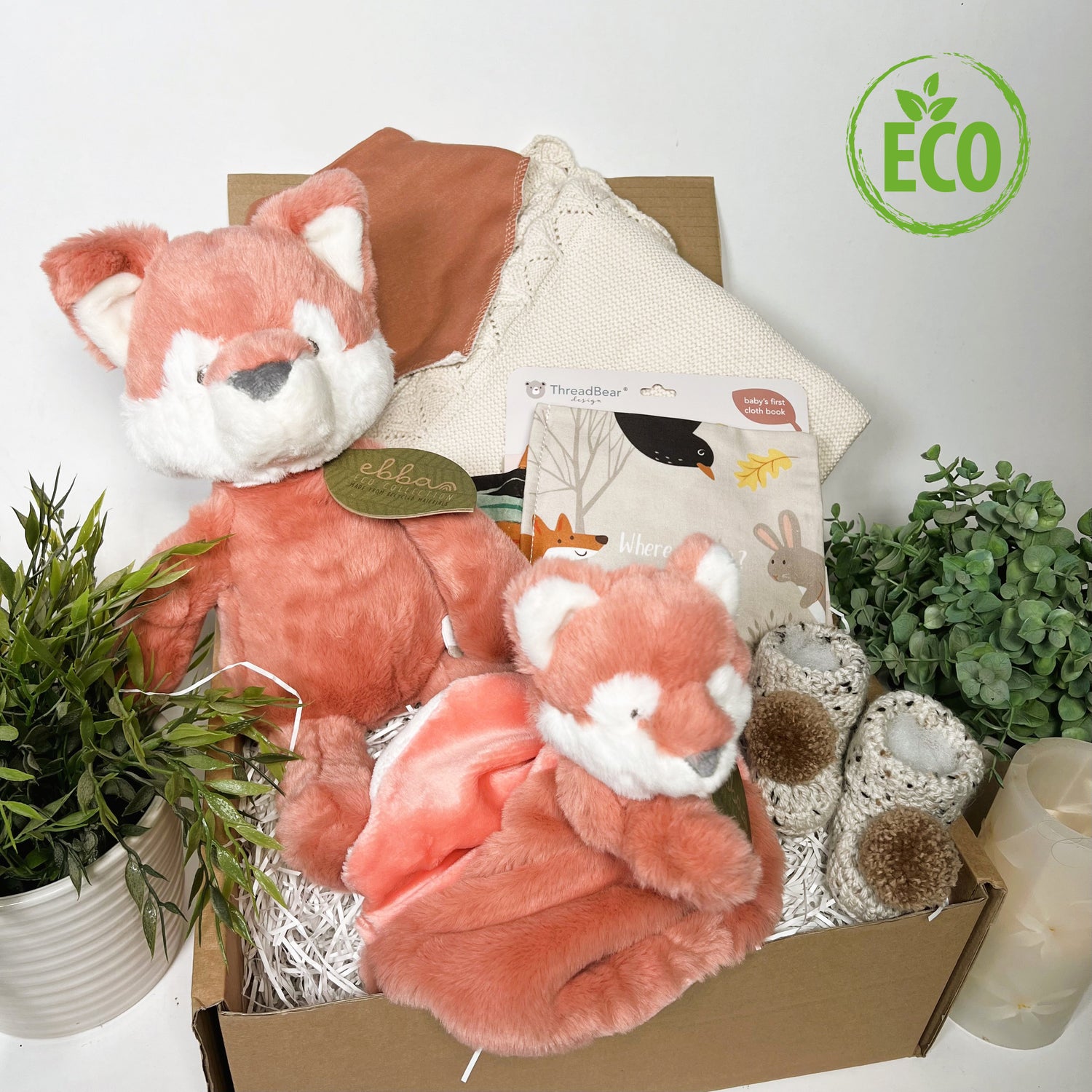 Neutral New Baby shower gift full of eco freindly items including a cotton baby blanket, a cloth baby book, an Ebba Fox soft toy and Ebba fox baby comforter, a cotton baby bib and a pair of crocheted baby booties with pompoms.
