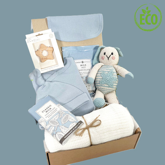 Eco friendly new baby boy gift icludes a blue baby sleepsuit with fold over cuffs and amatching baby knot hat and baby bib in a denim blue colour,  a white cotton cellular baby blanket, a pouch of Choc Affiar hot chocolate and a bar of milk chocolate, a Bambino star shaped wooded baby teether and a blue crochet bunny from Pebblechild.