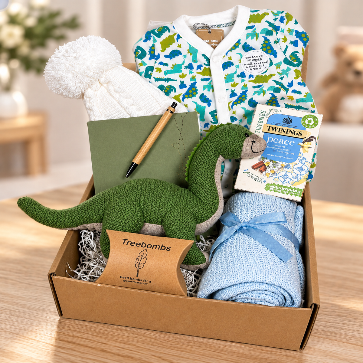 A baby gift hamper featuring a green knitted dinosaur plush toy, a white knit baby pom-pom hat, a light blue cellular baby blanket tied with a ribbon, a green notebook with a pen, a dinosaur print baby sleepsuit and a package of Twinings tea.