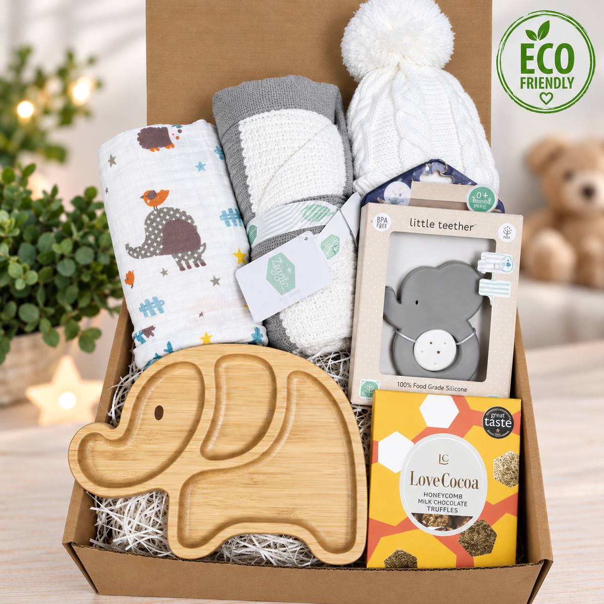 A curated eco friendly baby gift box filled with baby items, including patterned muslin swaddle, a knit baby hat with a pom-pom, a wooden elephant-shaped baby plate, a boxed elephant teether and a white cellular baby blanket with a grey trim and an 'ECO FRIENDLY' badge is shown in the top right.