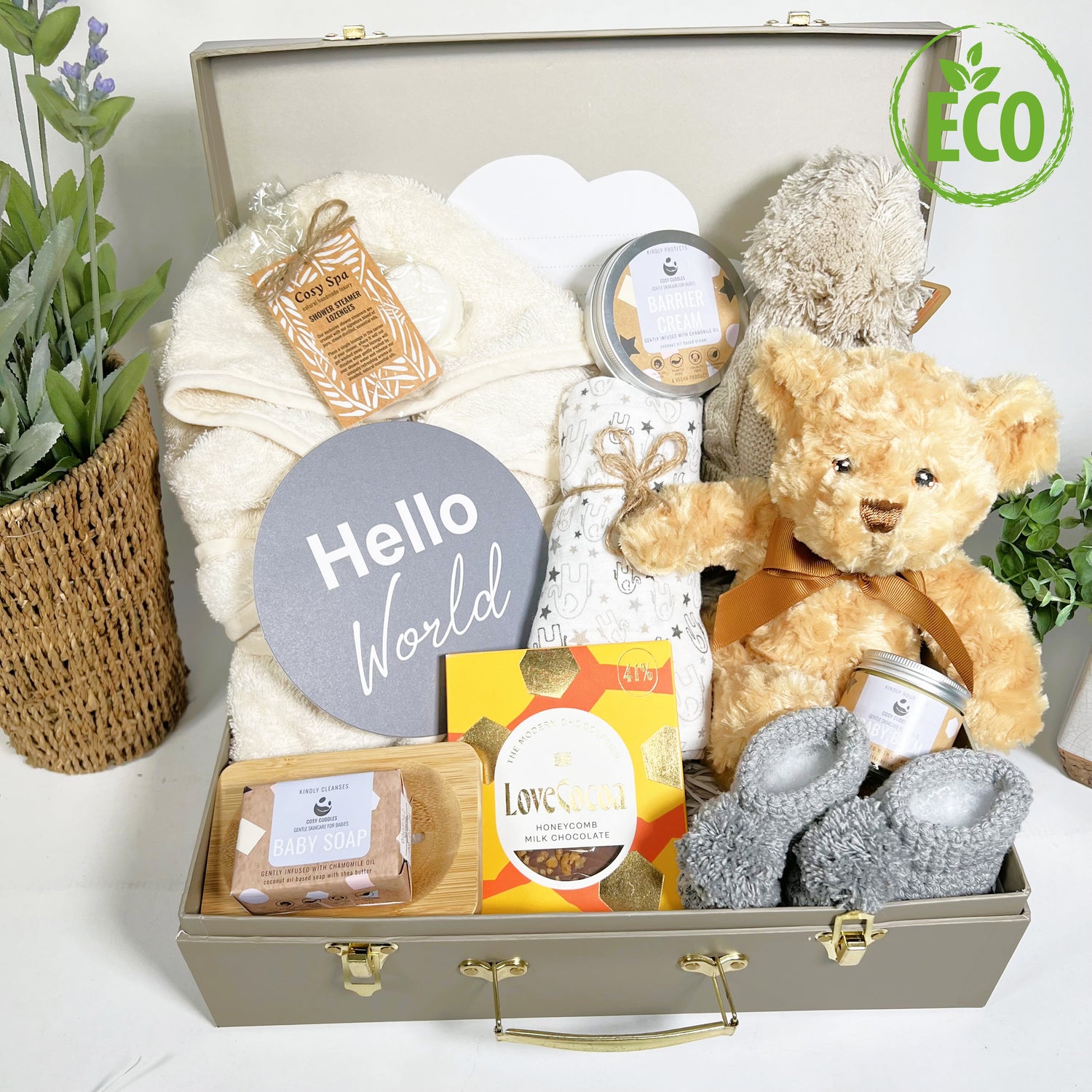 Gorgeous neutral new mummy and baby hamper containing eco friendly baby toiletries, a cotton baby dressing gown with hood, an eco friendly teddy bear baby toy, a baby pompom hat, a bar of chocolate and some shower spa steamers for Mummy, a pair of hand crocheted baby booties with pompoms,q muslin square and a "Hello World" plaque, all in a case