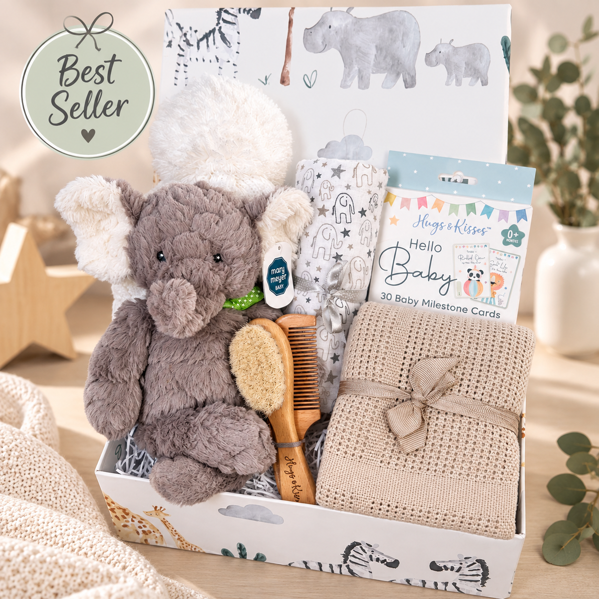 A baby gift basket set in a white box with animal illustrations, featuring a plush elephant toy, a beige knitted blanket tied with a ribbon, a wooden baby brush, a pack of Hello Baby milestone cards, and a Huggie Kit card. A green 'Best Seller' badge is in the top left, with a soft neutral background and decorative props.