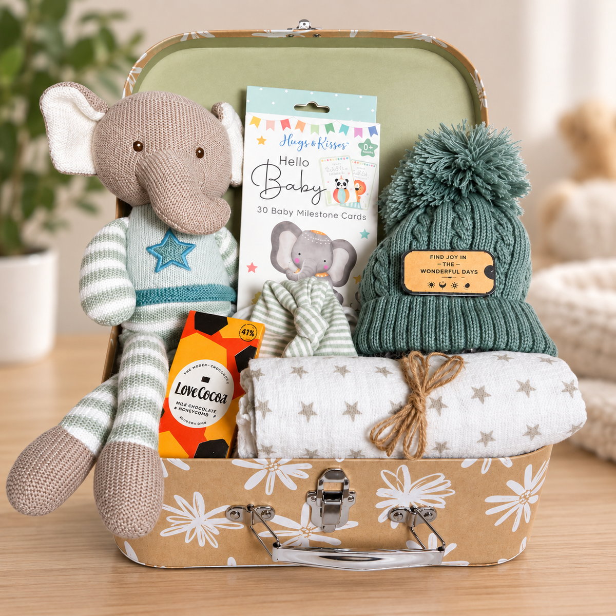 New baby hamper with a knitted elephant soft toy, a white muslin square with small star print, a green baby pompom hat, a white and taupe cotton baby knotty hat, a pack of baby milestone cards and some trets for mummy.