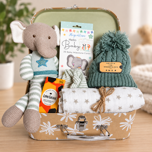 New baby hamper with a knitted elephant soft toy, a white muslin square with small star print, a green baby pompom hat, a white and taupe cotton baby knotty hat, a pack of baby milestone cards and some trets for mummy.
