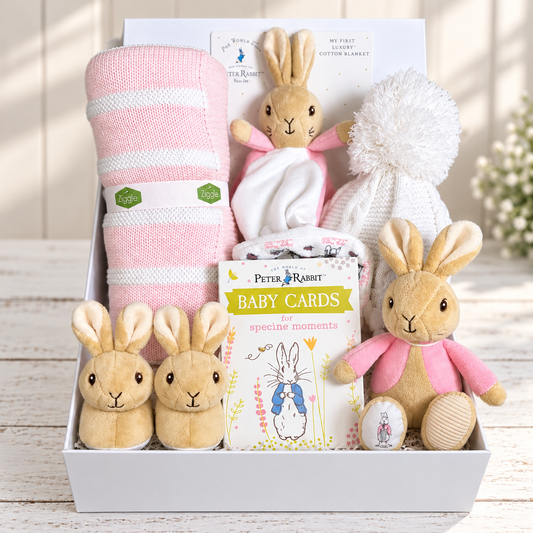 A curated Flopsey bunny baby gift hamper arranged in a white baby keepsake box, featuring a pink and white striped baby blanket, a white knit baby hat with a fluffy pom-pom, a plush Beatrix Potter rabbit toys, and baby comforter, a set of memorable moments cards and a pair of Peter Rabbit baby slippers.