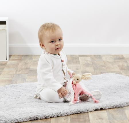 Baby sitting on a rug holding a Flopsey bunny comfort blanket plush toy in a room with a white wall.