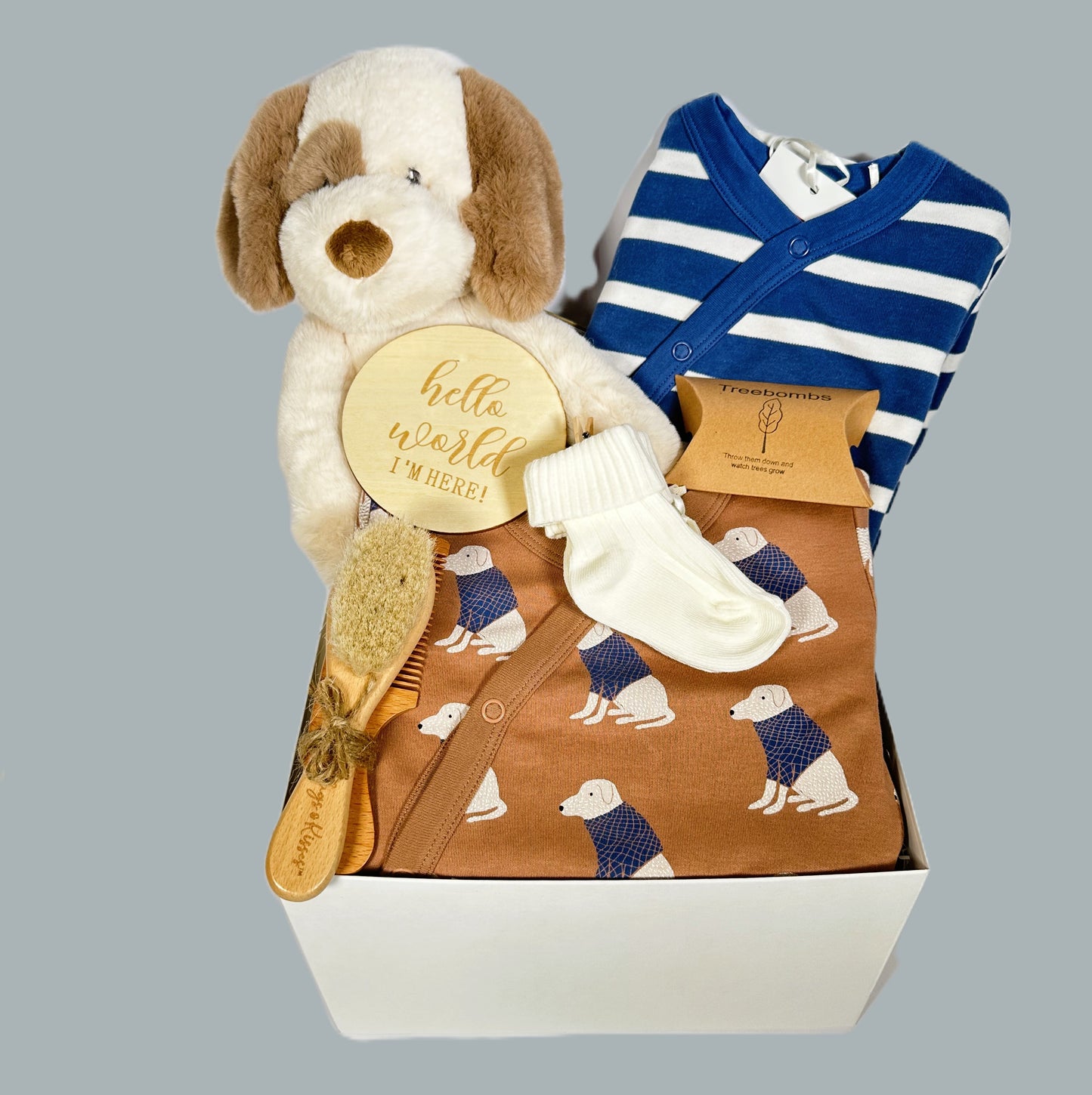 Baby hamper containing two organic cotton rompers with fold over cuffs and feet. One is a blue and white horizonal stripe and the other the same colours but with a labrador dog print. A baby brush and comb set and an Ebba Toddy dog soft baby toy.