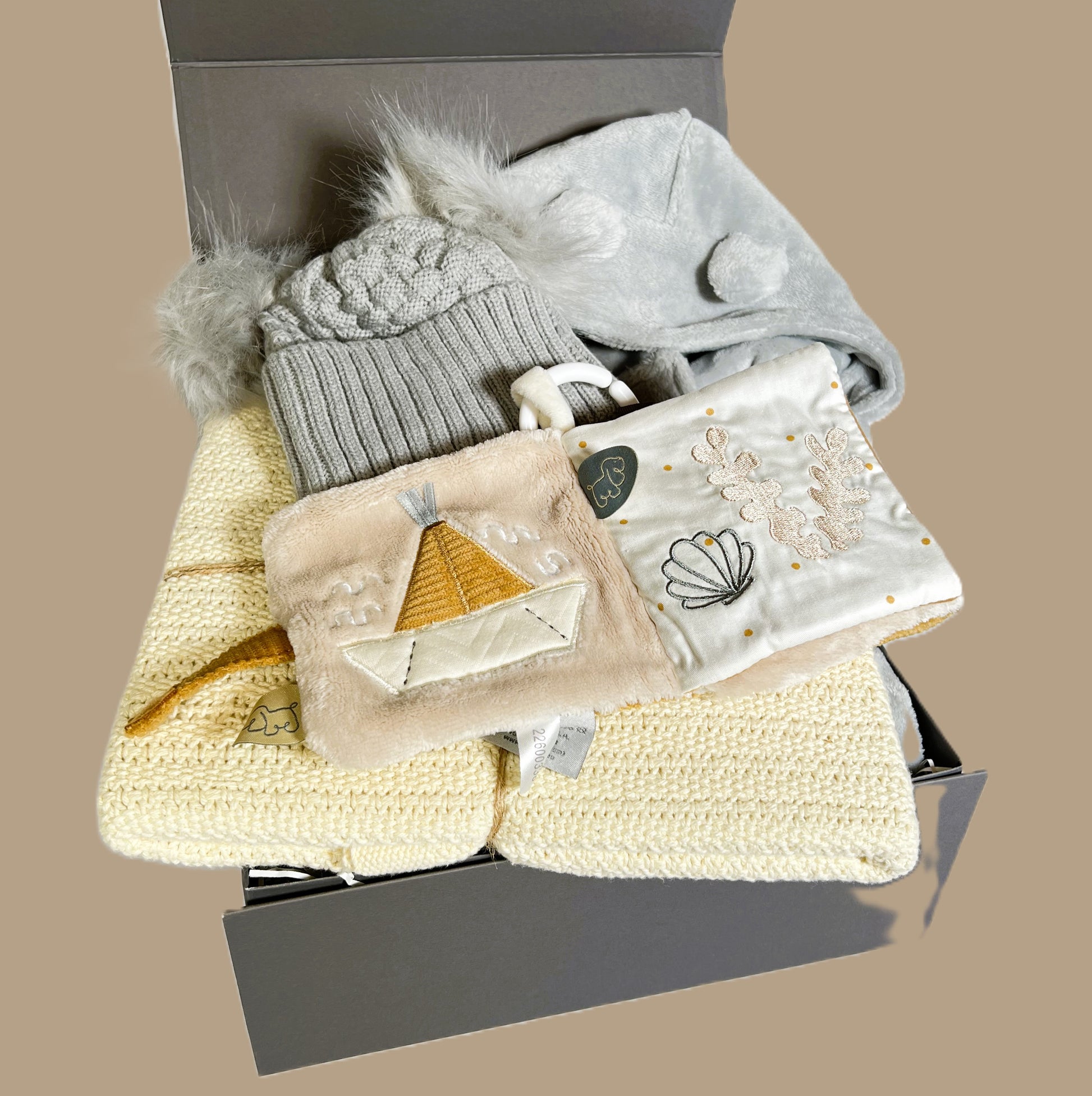 A baby hamper gift in a grey baby keepsake box containing a almond Hippychick cellular cotton baby blanket, a Nattou Charlie baby sensory book, a grey baby dressing gown and a baby pompom hat.