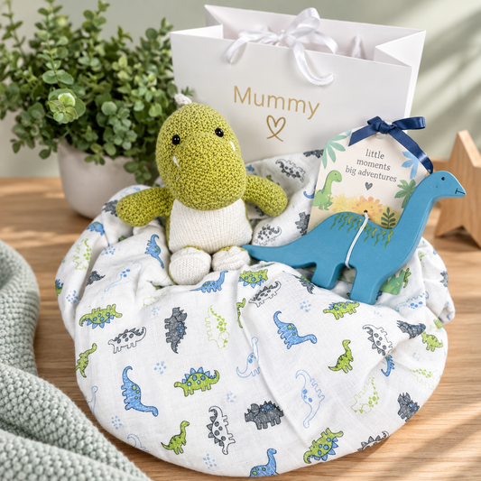 A  curate baby gift hamper featuring a white muslin swaddle blanket with a colourful dinosaur print, a yellow-green knitted dinosaur plush toy, a blue dinosaur-shaped natural rubber baby teether, a white gift bag printed with 'Mummy'.