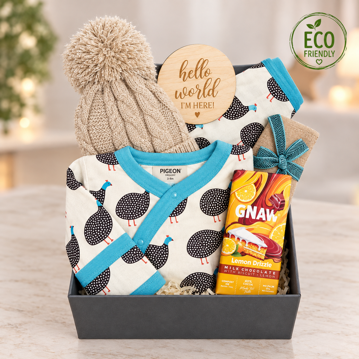 A new baby gift in a magnetic gift box containing an organic baby sleepsuit in a Guinea fowl print with matching reversible baby dribble bib, A bar of Gnaw chocolate, a taupe baby muslin and a taupe baby pompom hat.