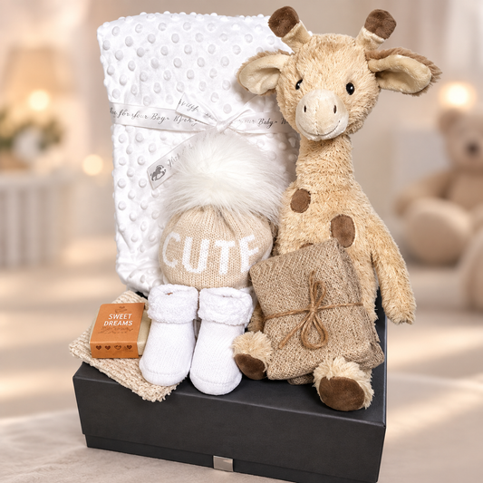 A baby gift set arranged in a baby keepsake  box, featuring a plush giraffe toy, a white bubble baby blanket, a beige knit hat with white faux-fur trim and the word 'CUTE' on it, white baby socks, a bar of essential oils soap  reading 'Sweet Dreams', and a soap bag tied with string, photographed in a softly lit room.