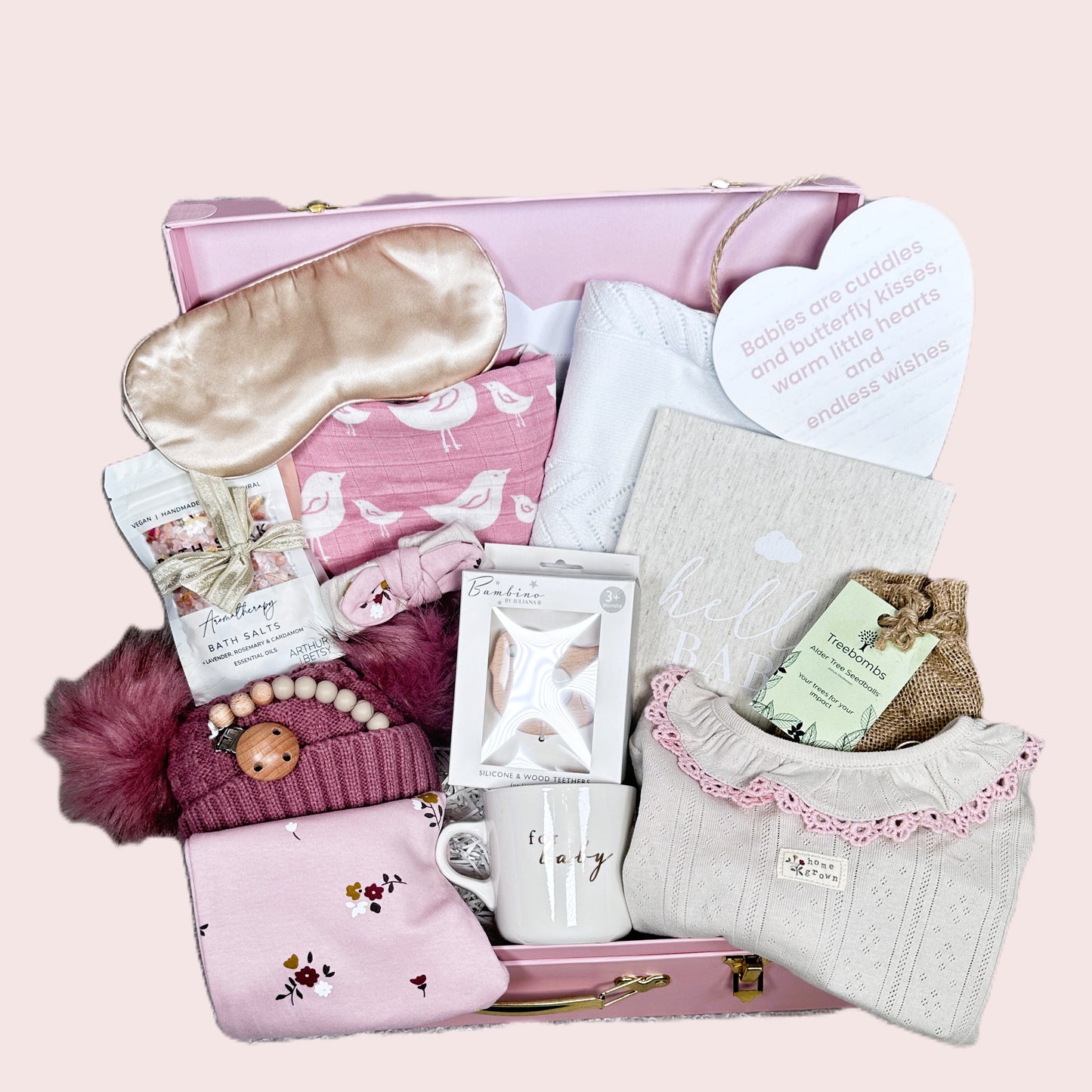 baby hamper for a girl with various items including a sleep mask, mug, and babywear on a pink background