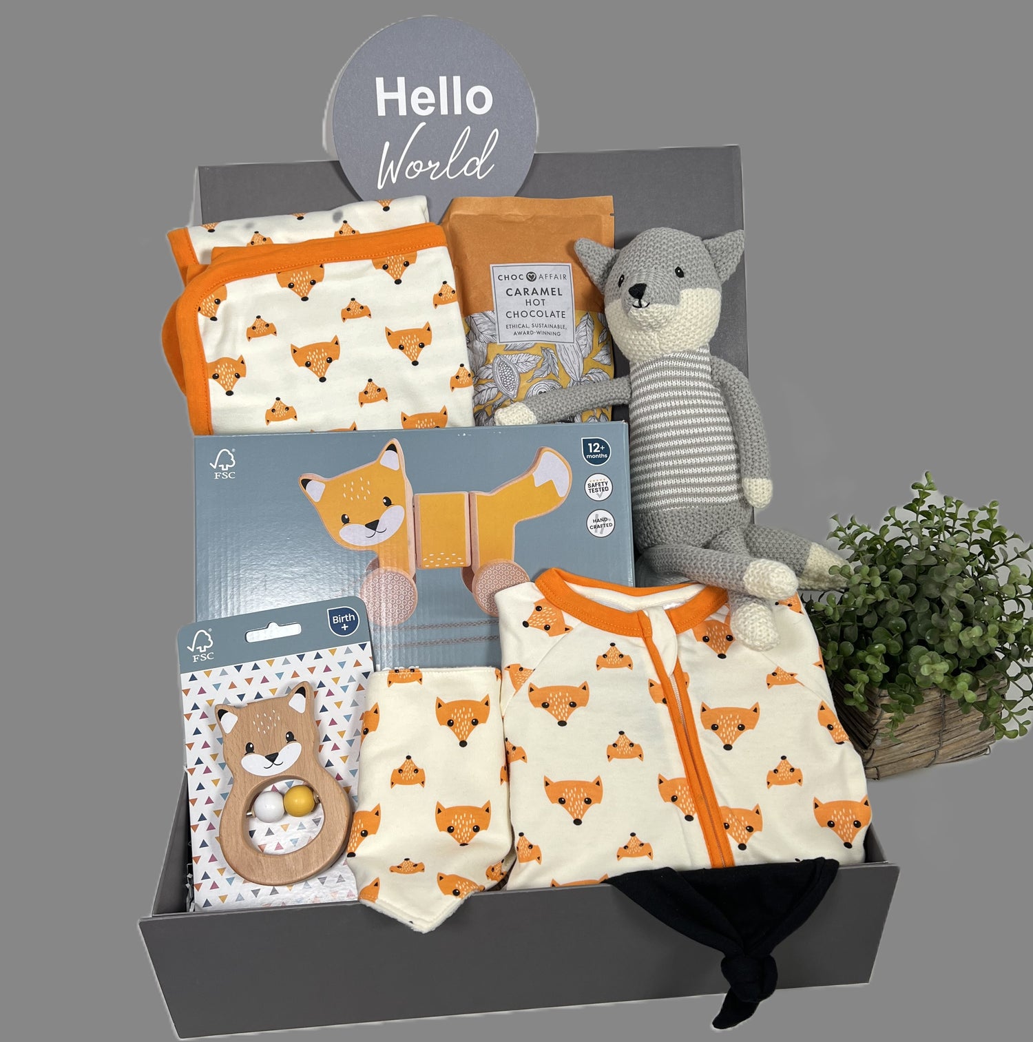 neutral new baby gift fox themed baby hamper with cotton zipped sleepsuit, matching hooded baby blanket and dribble bib, a fox soft toy, a wooden FSC pull along fox toy, a FSC fox baby rattle, a black baby knot hat and a pack of Choc Affair caramel hot chocolate.
