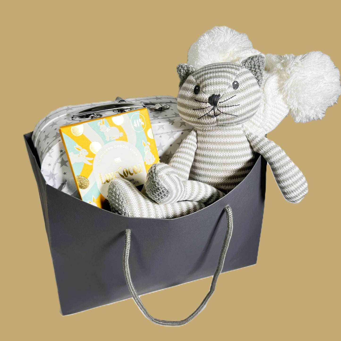 Neutral new baby gift in a bag containing a grey and white stripped knitted cat toy, a white baby pompom hat, a bar of Loce Cocao choclate, a kitty print baby layette set and a white storage suitcase with grey star print.