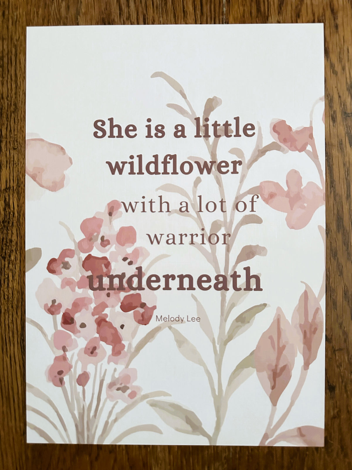 An A4  art print featuring a quote by Melody Lee, 'She is a little wildflower with a lot of warrior underneath,' printed in dark brown text over a watercolour floral design in dusty pink and muted green on a white background. The print is displayed flat on a dark wood surface.