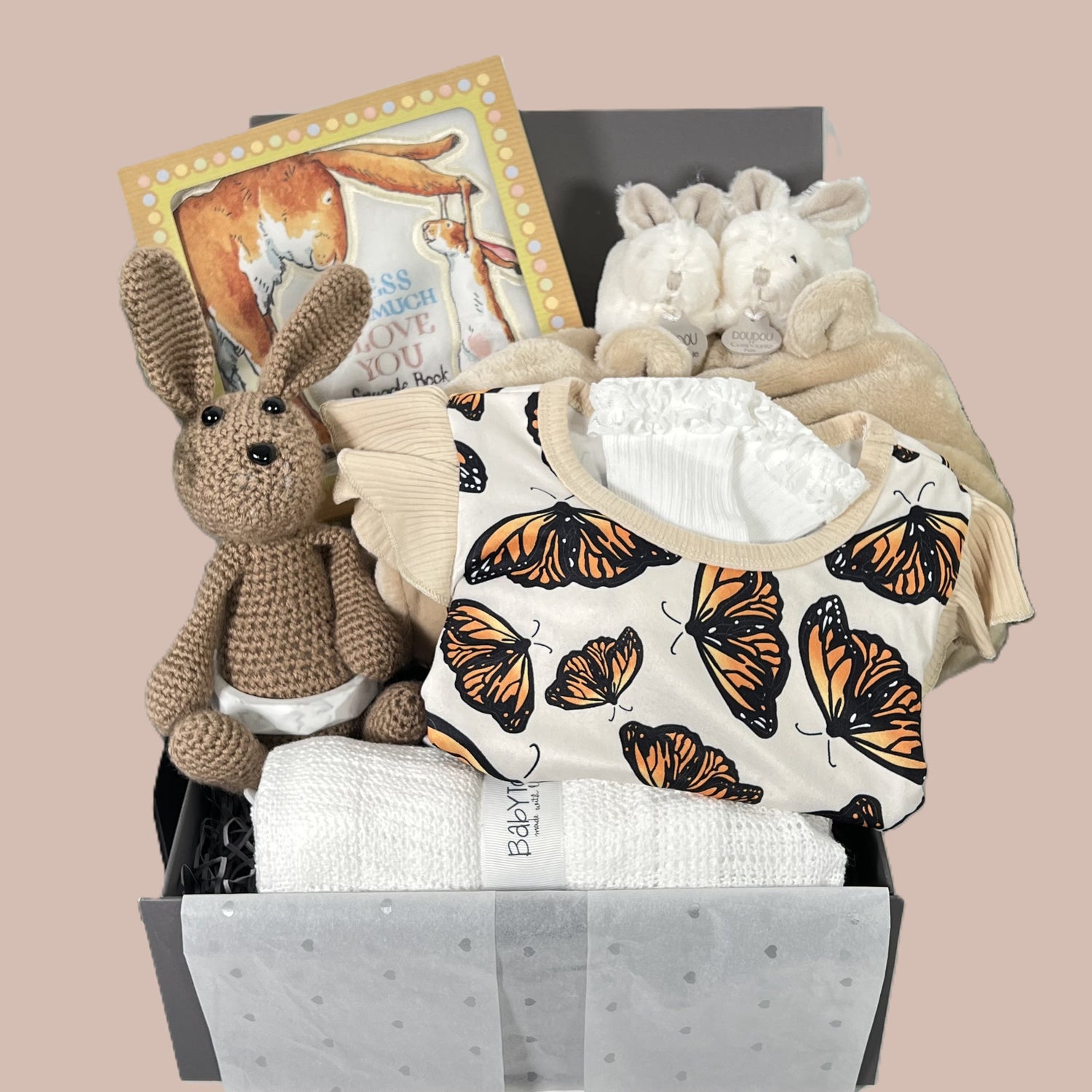 New baby girl hamper gift box with a butterfly romper, a baby dressing gown , a pair of Doudou Et Champagnie baby slippers, a Guess how much I love you snuggle soft baby book, a white cotton cellular baby blanket and a crocheted bunny baby.