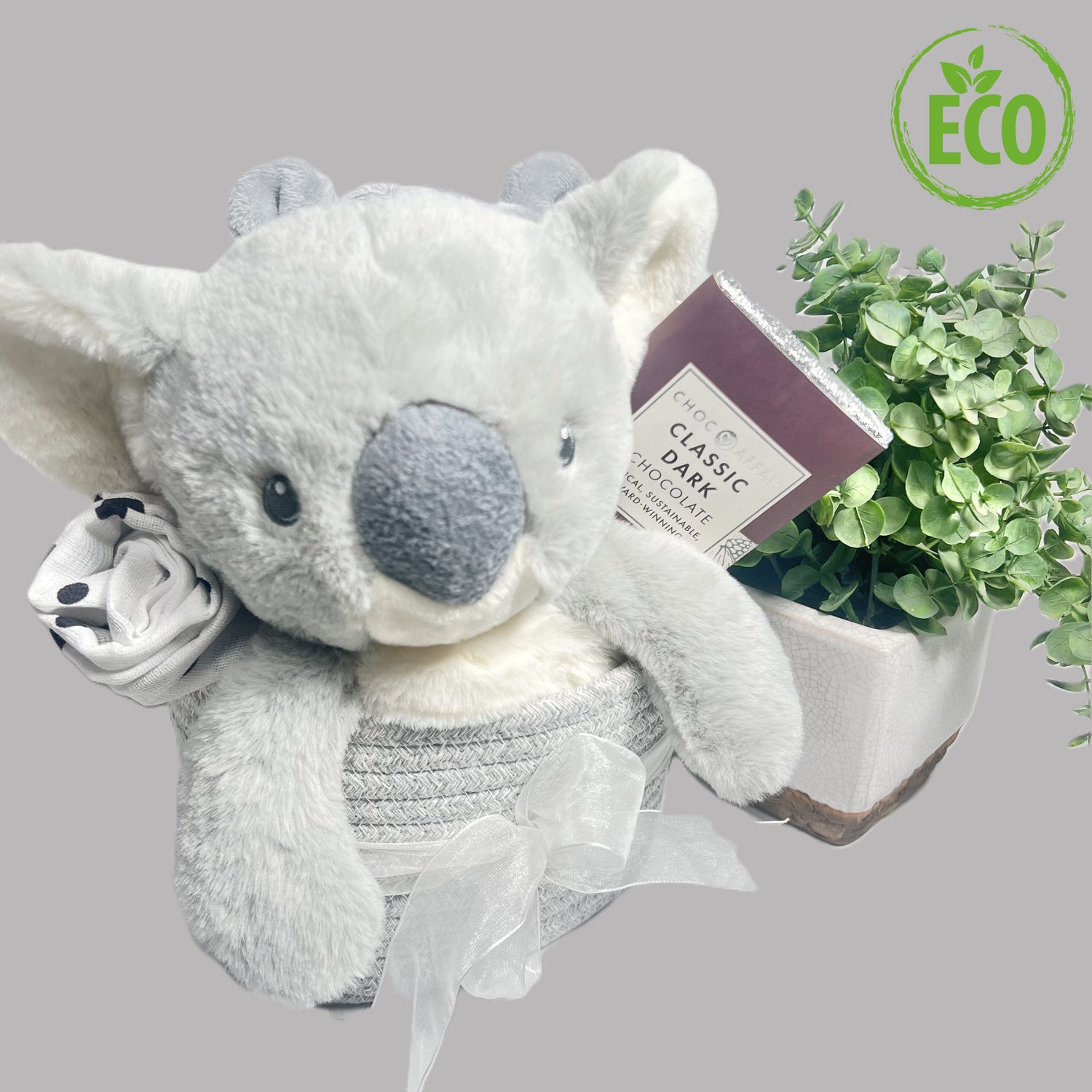 A mummy and baby hamper basket in a rope storage caddy containing a Keeleco 25cm Koala bear soft baby toy, a white baby dressing gown , 90gm bar of Choc Affair milk or oatmilkchocolate and a white and black large baby muslin square.