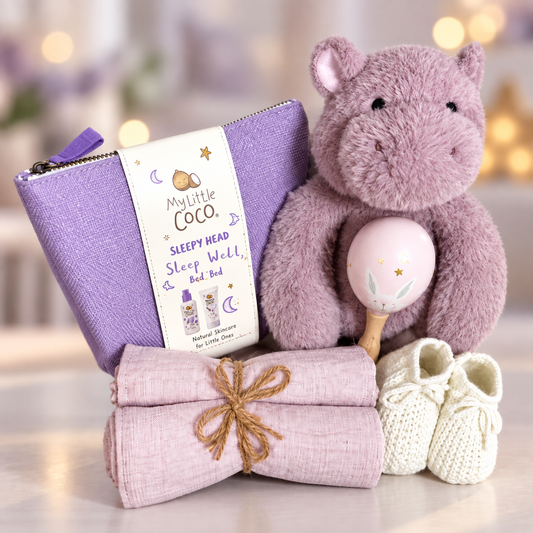 A baby gift set featuring a lavender zip pouch with a 'My Little Coco Sleepy Head' paper band, a folded light pink baby muslins tied with twine, a plush lavender hippo toy holding a pink maraca, and a pair of white knit baby booties, arranged together on a light surface with a soft blurred background.