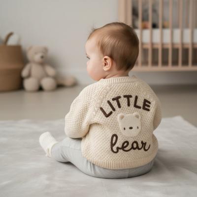 Baby wearing a 'Little Bear' baby cardigan in cream, sitting on a bed in a nursery.
