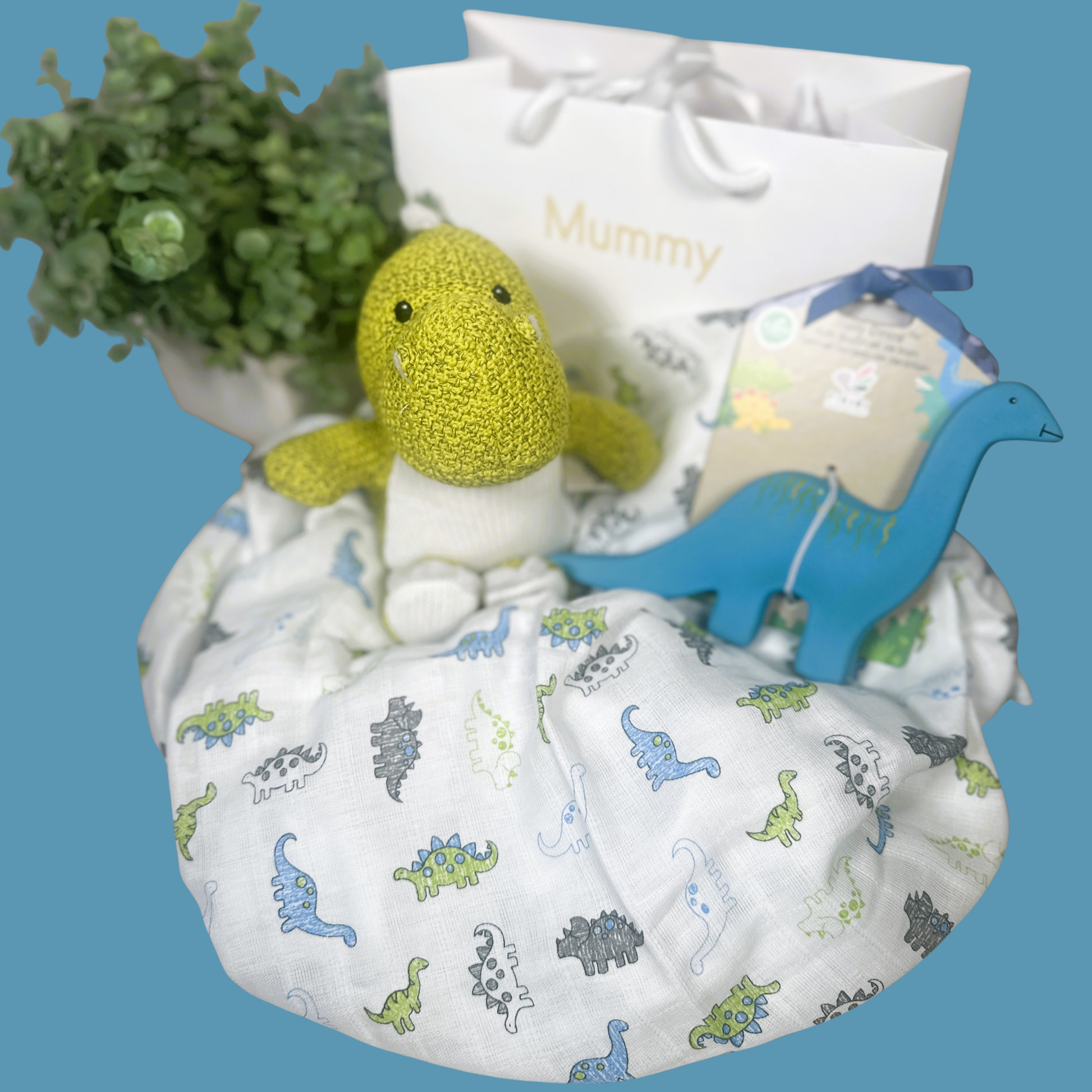 A baby gift set featuring a white muslin swaddle blanket with a colorful dinosaur print, a yellow-green knitted dinosaur plush toy, a blue dinosaur-shaped toy, a white gift bag printed with 'Mummy', and a natural rubber baby teether shaped like a dinosaur arranged on a solid blue background.