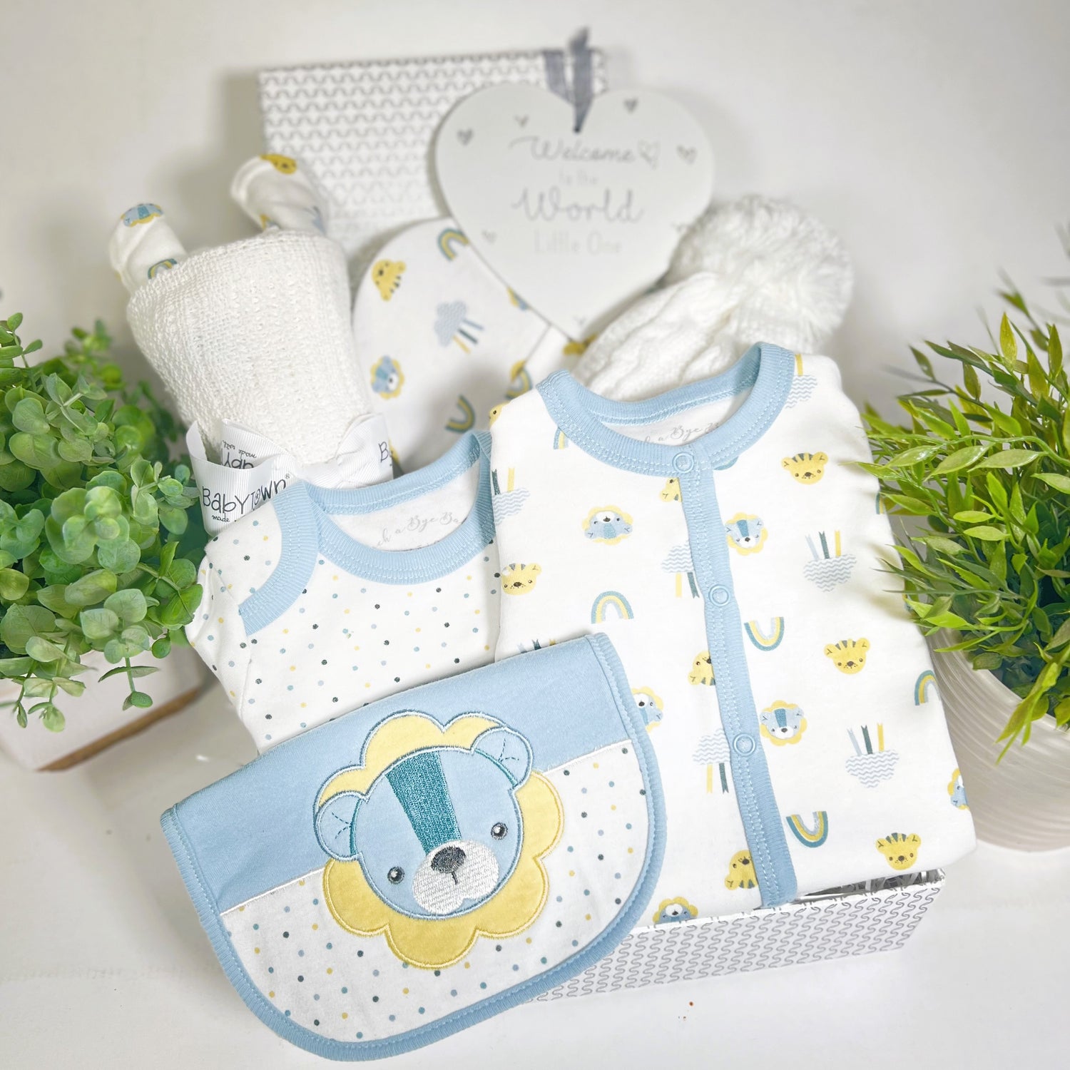 A new baby boy baby shower gift in a silver  and white baby keepsake box containing a cotton layette set with blue and yellow lion print, a white cotton cellular baby blanket, a baby pompom hat and a grey and white heart shaped nursery sign.