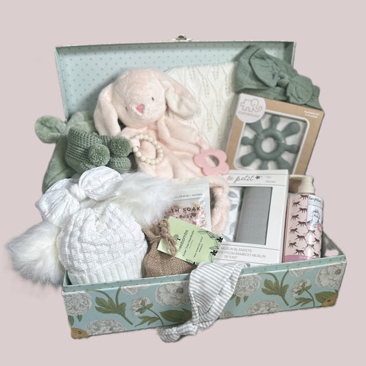 Gift box with baby items including a plush toy, hat, and soap on a light pink background