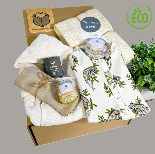 Luxury Eco Friendly unisex new baby hamper containing a almond coloured cotton cellular baby blanket, a GOTS certified cotton print baby romper with a green and black print of a Lesser Antillean Iguana, a pot of natural baby baby and a tin of natural baby barrier cream, a muslin square in brown, a natural cotton baby bathrobe a Unnaaty soya candle and a small grey and white baby photography disc which read "I'm new here" and "Little and loved" on the other side