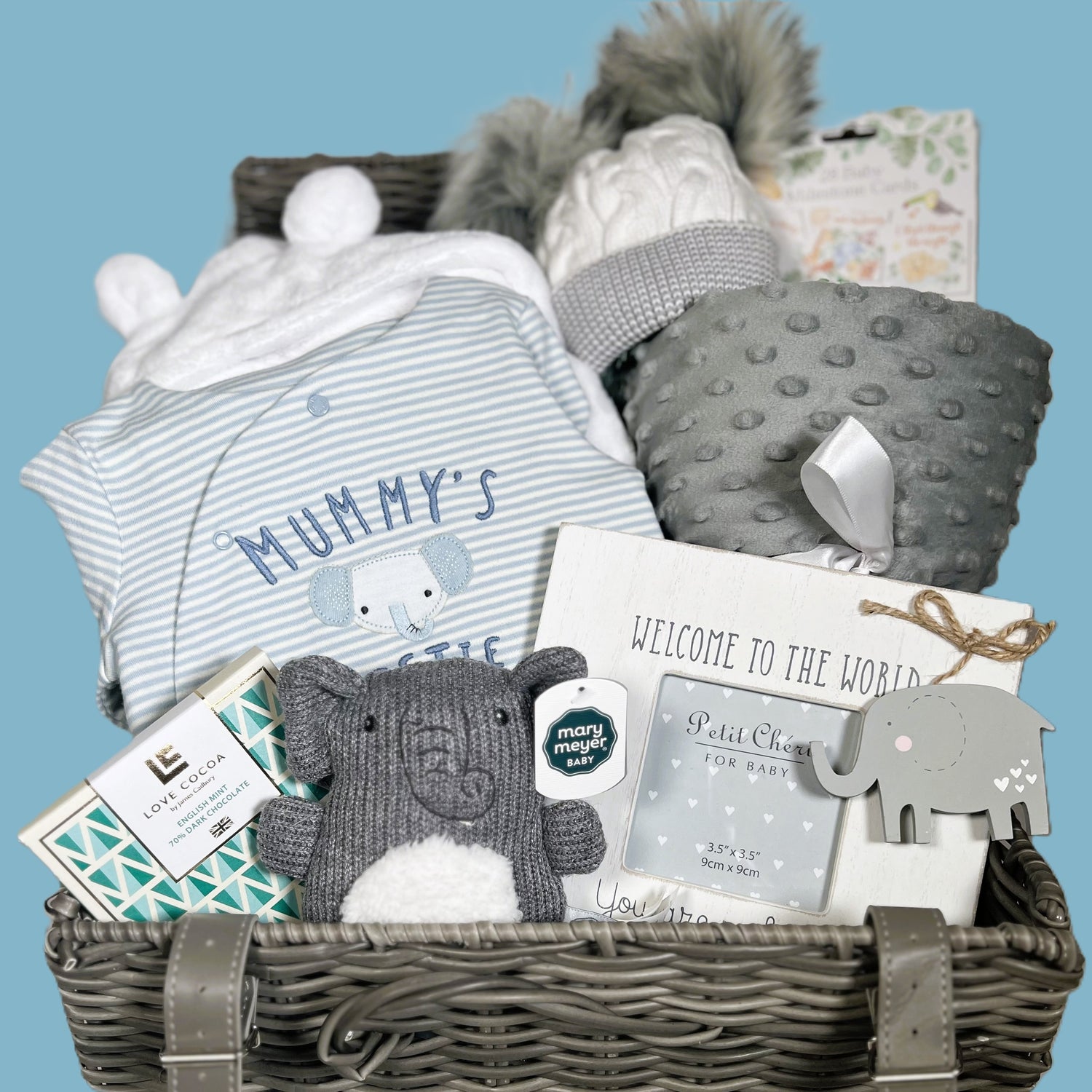 A baby shower gift in a woven baby keepsake case containing q white hooded baby dressing gown, a cotton blue and white striped baby sleep suit with an embroidered elephant face and the words "mummy's bestie", a grey sherpa baby blanket, a grey and white baby pompom hat,a mary Myer knitted elephant baby rattle, a baby photo frame and a bar of Love Cocoa chocolate