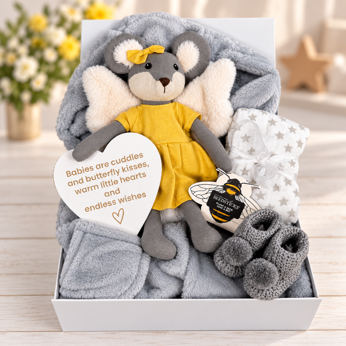 A baby gift box set containing a plush grey mouse toy with white angel wings and a yellow dress, a soft grey baby robe, a white star-patterned baby muslin, a pair of grey crocheted baby booties, and a heart-shaped nursery plaque that reads 'Babies are cuddles and butterfly kisses, warm little hearts and endless wishes'. Bee seedsticks