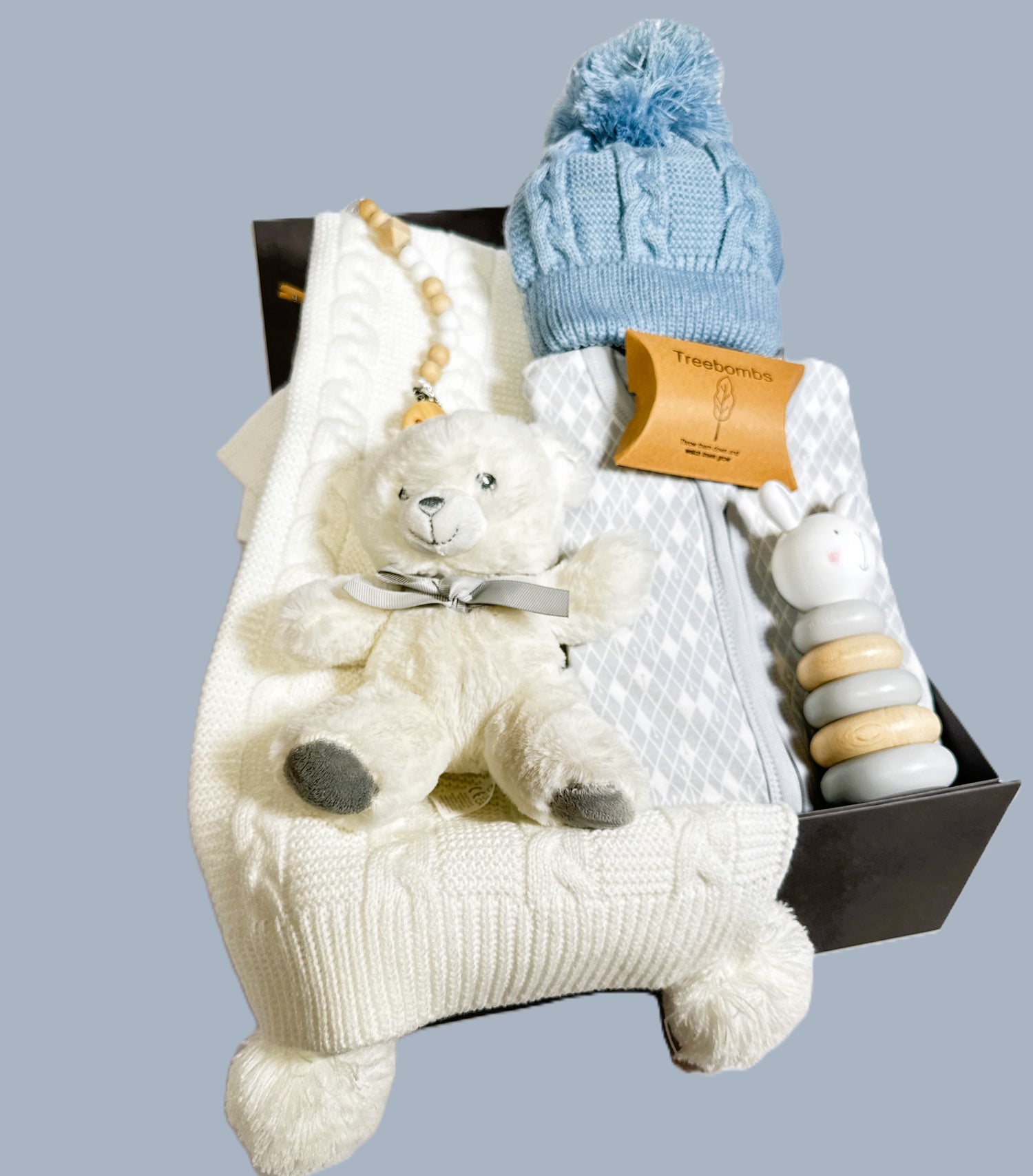 Baby boy hamper in a grey baby keepsake case with a zipped baby sleepsuit, a blue baby pompom hat, a dummy clip, white and grey teddy bear, a white baby blanket and a Bambino stacking baby toy.