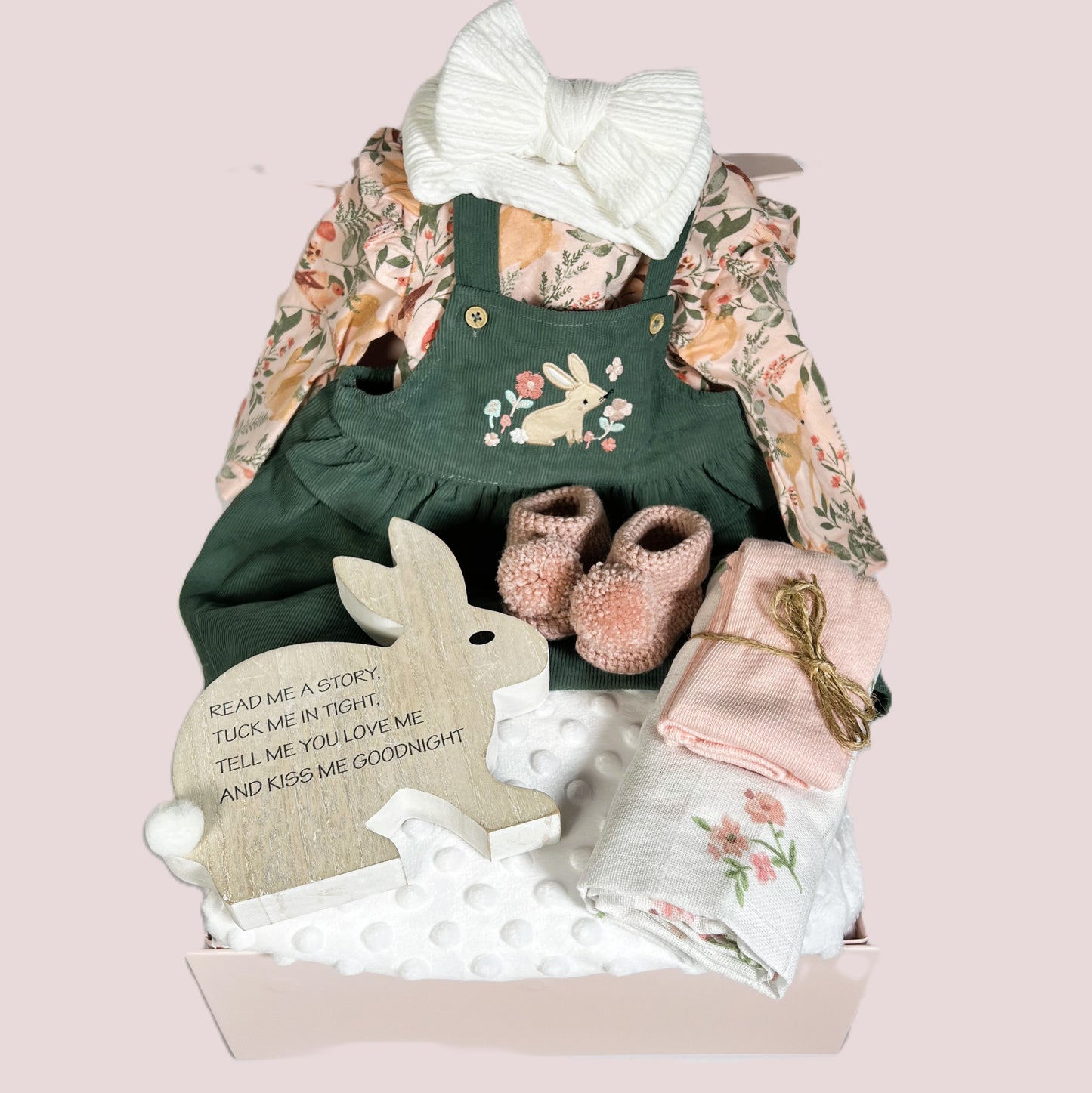 A new baby girl hamper with a corduroy pinafore dress and body suit with long sleeves and matching peach coloured baby tights. A pair of crocheted baby booties with pom poms. A white baby bubble blanket a, wood nursery ornament and a floral print baby muslin square.