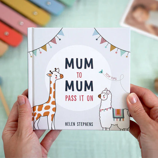 Book titled 'Mum to Mum Pass It On' by Helen Stephens held by hands with colorful objects in the background.