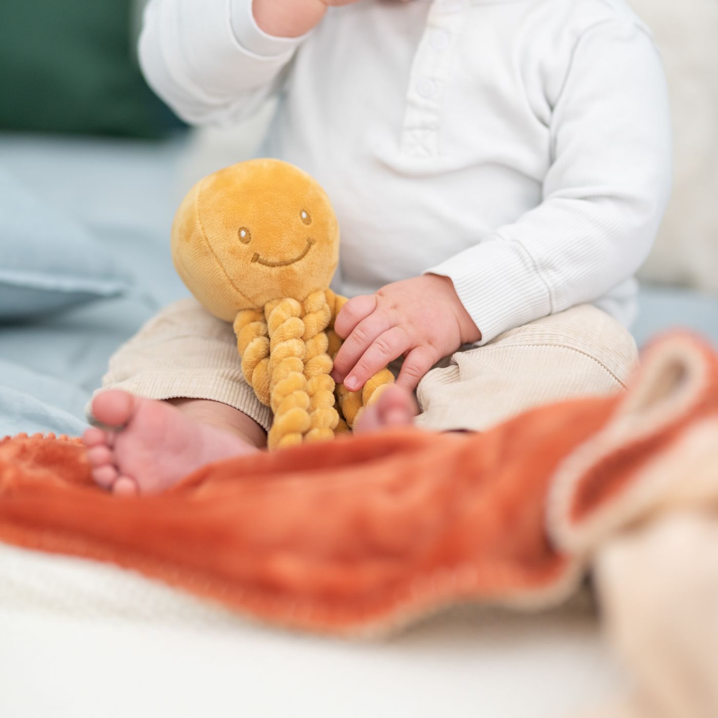 Child holding a Nattou Pui Pui Ochre with a smiley face, sitting on a couch with an orange blanket.