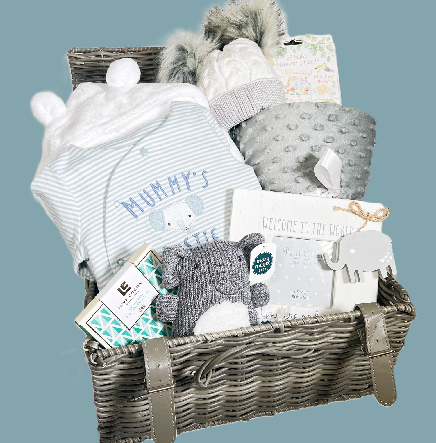 A baby shower gift in a woven baby keepsake case containing q white hooded baby dressing gown, a cotton blue and white striped baby sleep suit with an embroidered elephant face and the words "mummy's bestie", a grey sherpa baby blanket, a grey and white baby pompom hat, a Mary Myer knitted elephant baby rattle, a baby photo frame and a bar of Love Cocoa chocolate