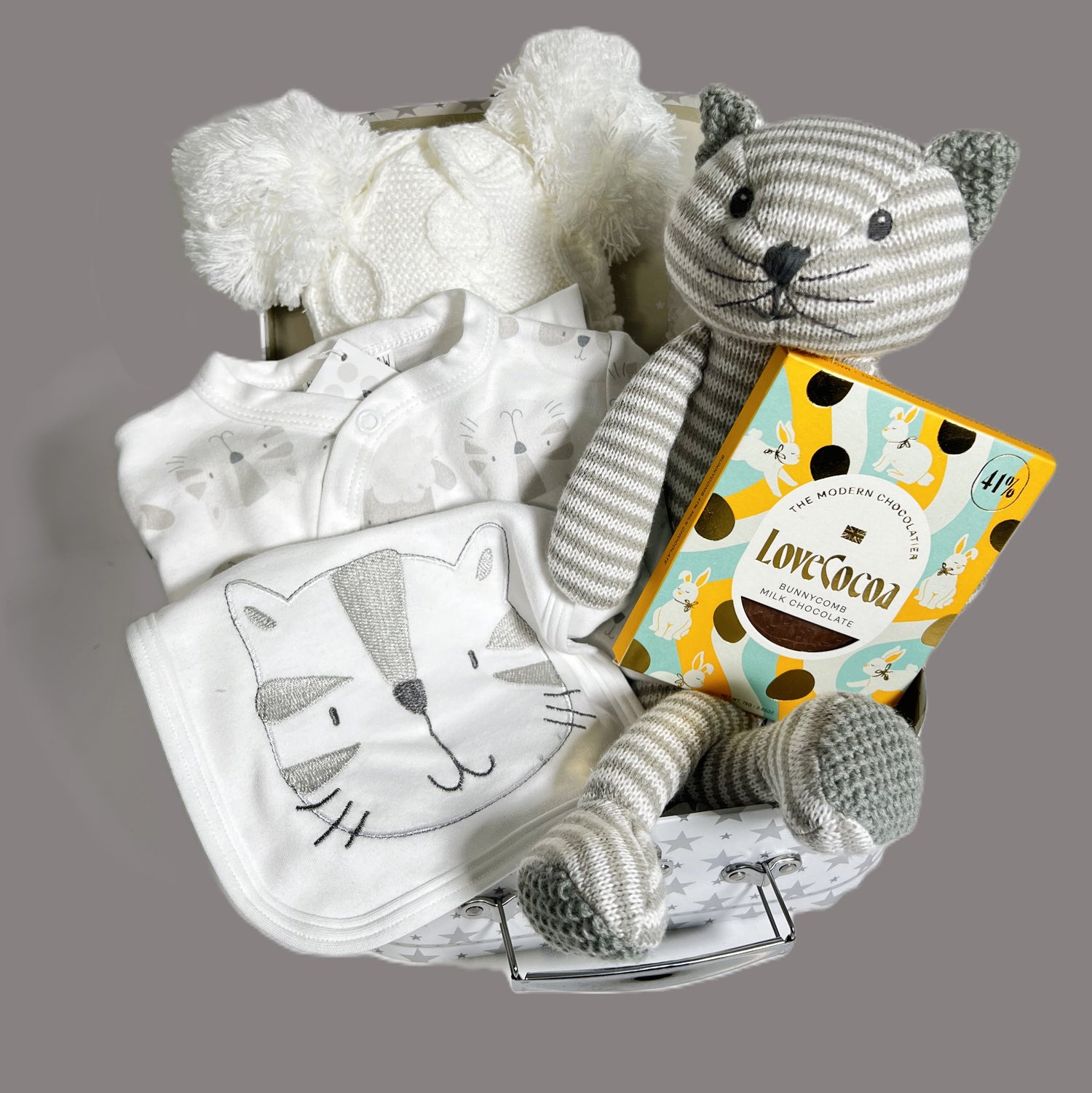 Neutral new baby gift in a bag containing a grey and white stripped knitted cat toy, a white baby pompom hat, a bar of Loce Cocao choclate, a kitty print baby layette set and a white storage suitcase with grey star print.