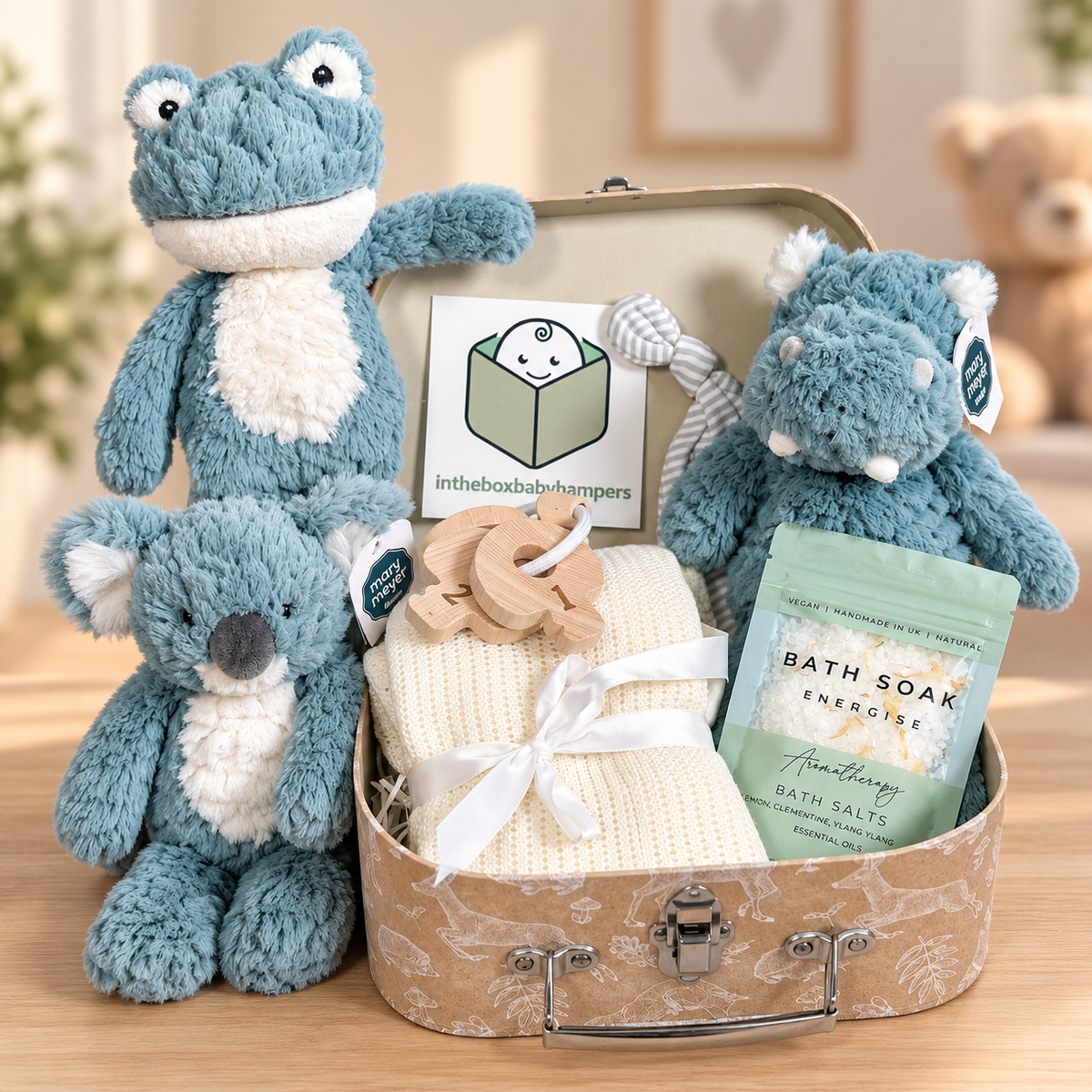 A plush toy gift set arranged in a vintage-style baby keepsake case on a wooden surface. The set includes a large blue frog plush, or smaller blue koala plush or a blue hippo plush, a cream knit baby blanket tied with a white ribbon, a wooden teething ring, a mint green bath soak pouch, and a small branded card.