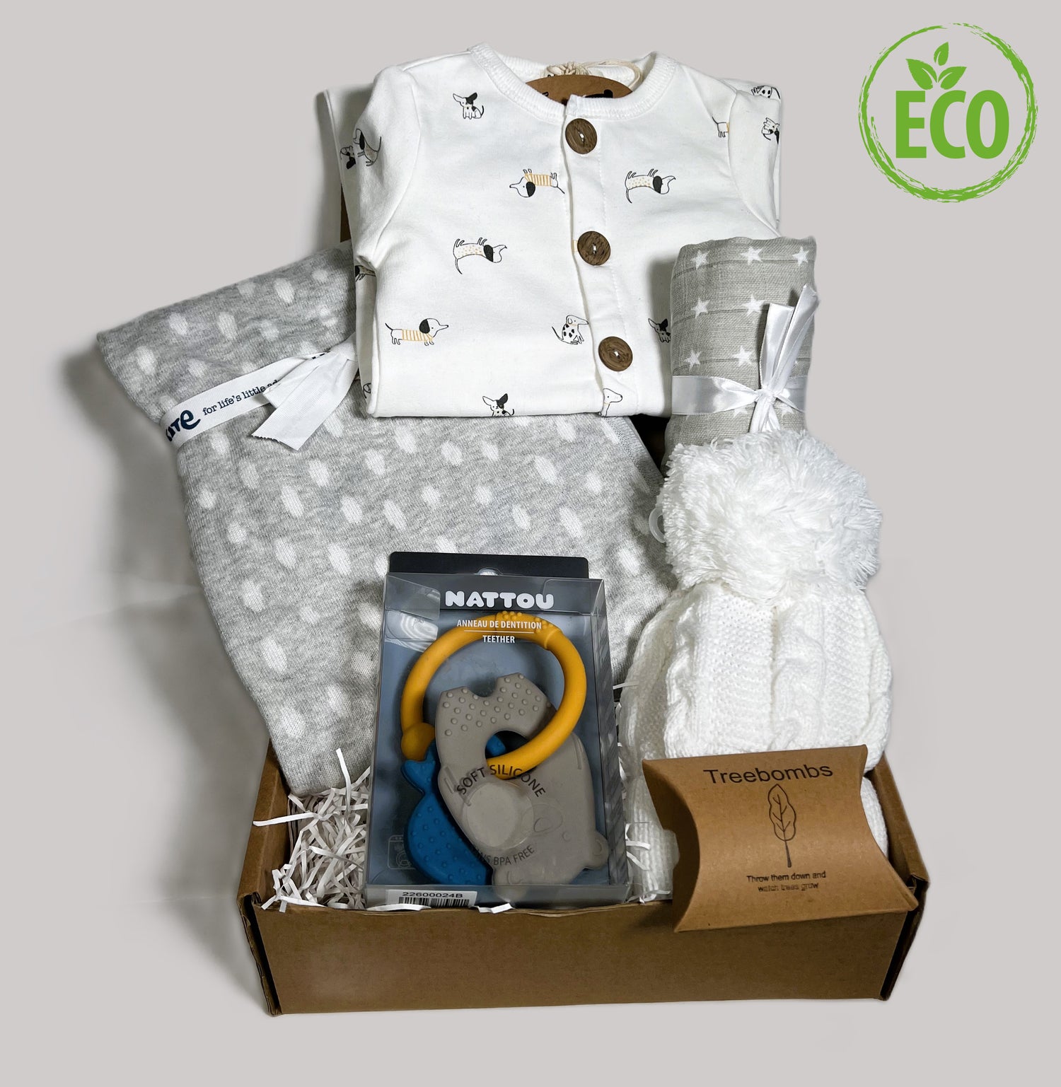 A neutral new baby gift containing a cotton button up romper with a sausage dog print, an organic cotton grey blanket with white oval print, a nNattou silicone baby teething toy, a grey ad white star print baby muslin and a white baby pompom hat.