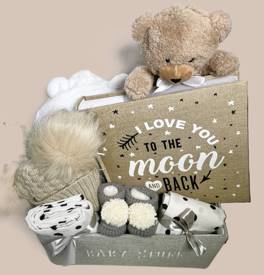 Unisex baby gift hamper in A "baby stuff"tote containing a "love you to the moon and back large baby photo album in neutral with white and black text, two baby muslin, a biscuit coloured baby pom pom hat a white baby dressing gown a Breon teddy bear soft baby toy and a pair of hand crocheted baby