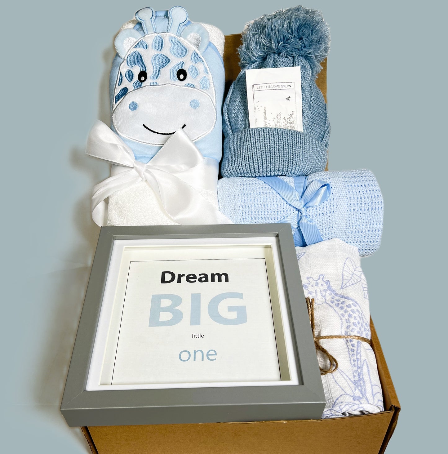 Blue baby shower gift containing a framed "Dream big little one " nursery print, a blue baby pompom hat, a blue and white hooded baby towel with an embroidered giraffe face, a blue cotton cellular baby blanket and a blue and white baby muslin.