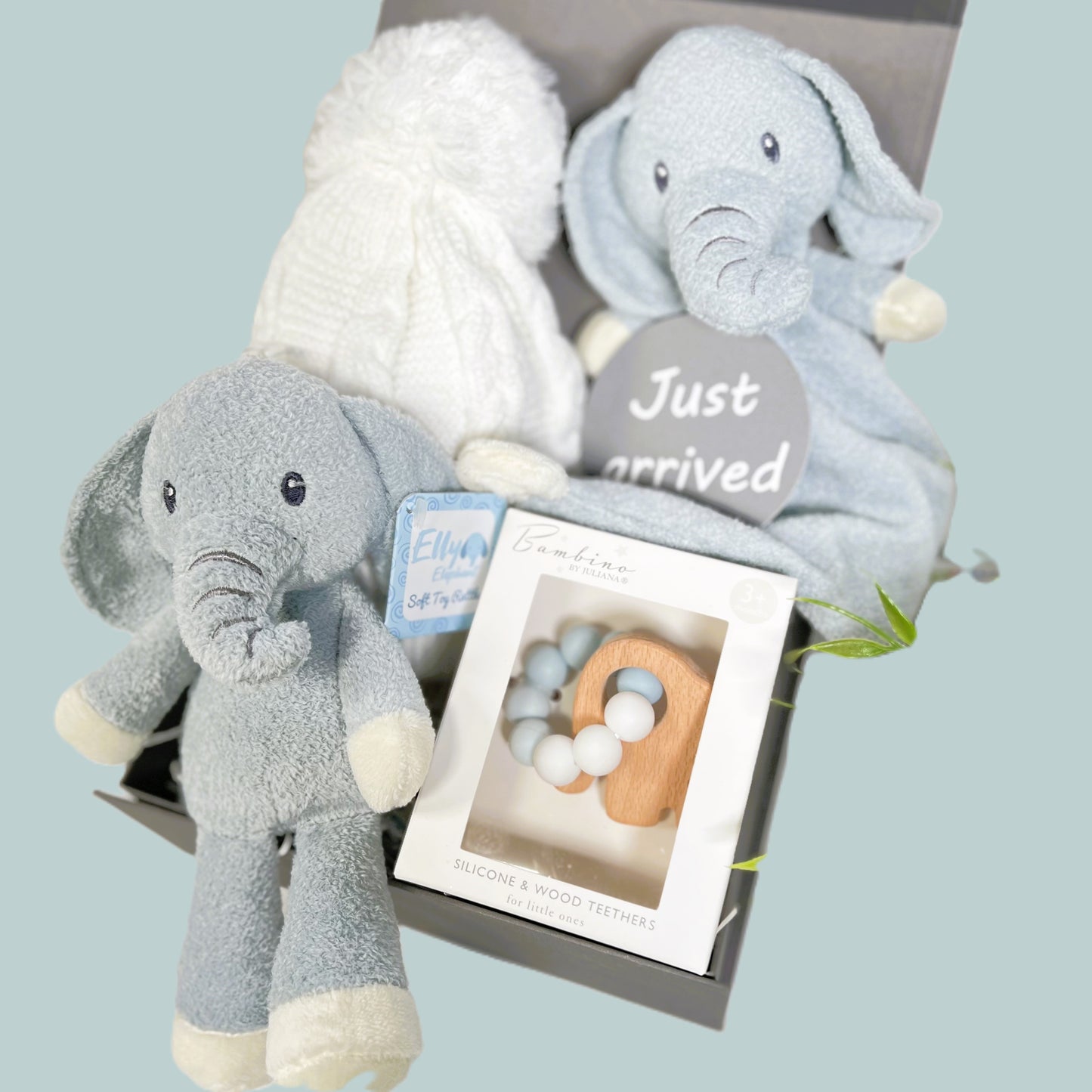 Blue and white new baby boy hamper contained in a grey baby keepsake box, a Blue and white elephant rattle toy with embroidered eyes and trunk and a matching Ely elephant baby blankie comforter. A white baby pompom hat made from recycled plastics, a "Just arrived" baby photography disc and a Bambino silicone and beech wood baby teether in blue and white with a wooden elephant shaped teether.
