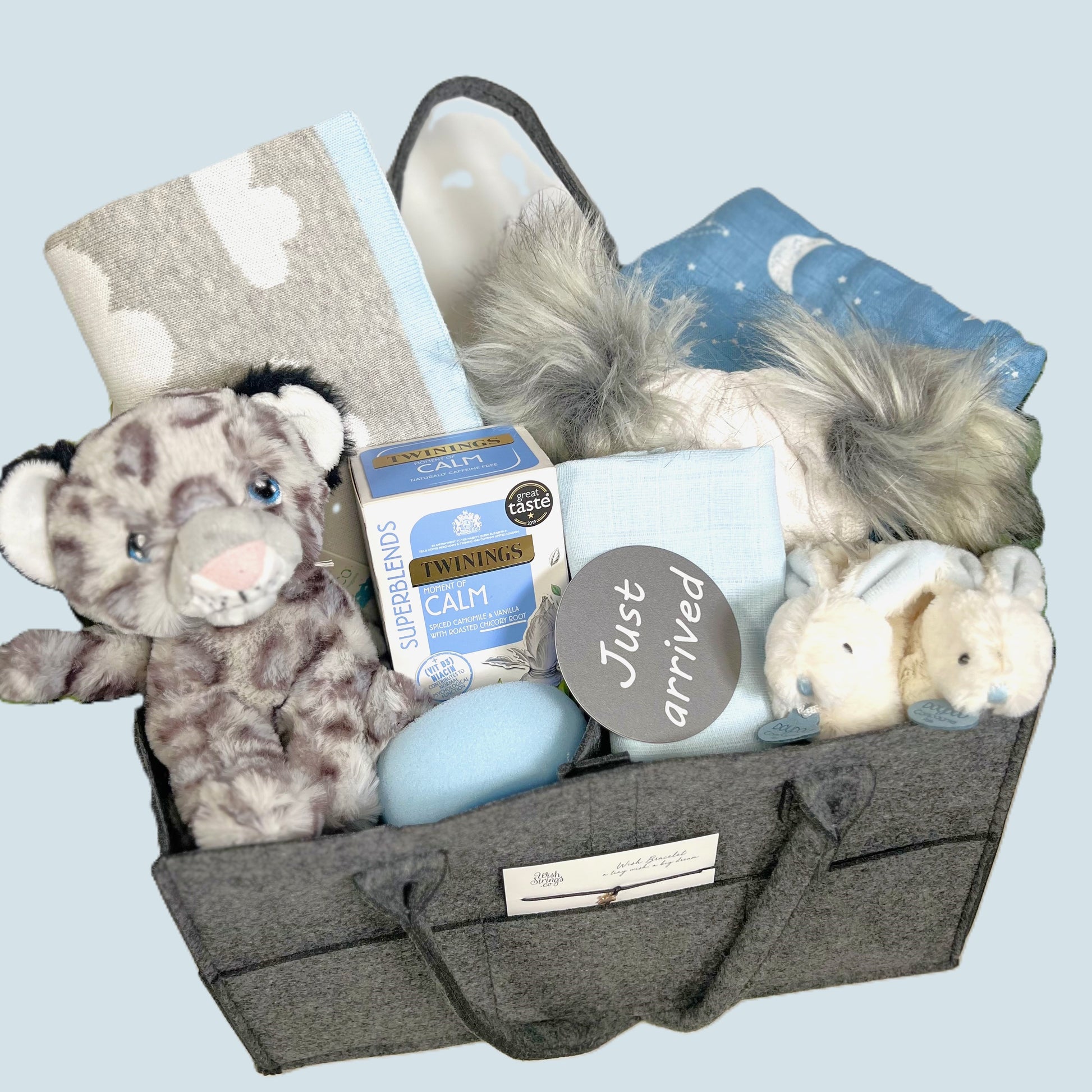 A New Baby Boy gift of a dark grey felt nappy caddy containing a cotton baby blanket in grey with a white coloud pattern and sky blue trim, a large blue baby muslin with a white print of planets, a Kelleco snowlepoard soft baby toy, a pair of Doudou Compagnie bunny baby slippers in white and blue, a grey and white baby pompom hat with two fluffy grey pompoms, a packet of Twining Calm teabags, a wish string and various plastic free baby toiletries.