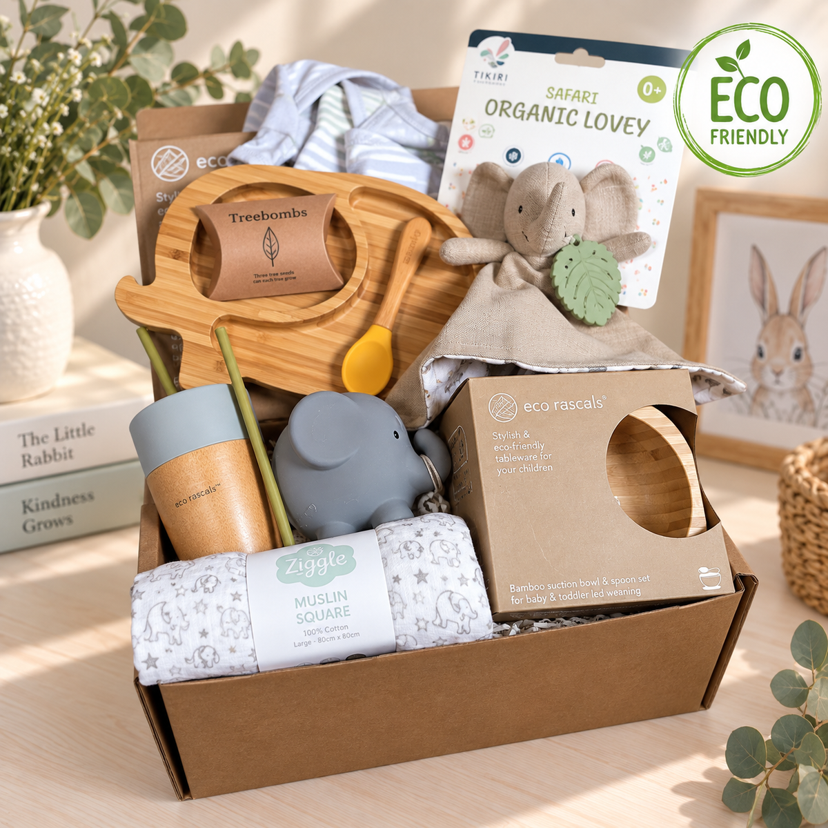 A curated new baby gift box filled with eco-friendly products including a bamboo baby plate, a muslin square blanket, a cotton elephant baby comforter, a bamboo baby cup