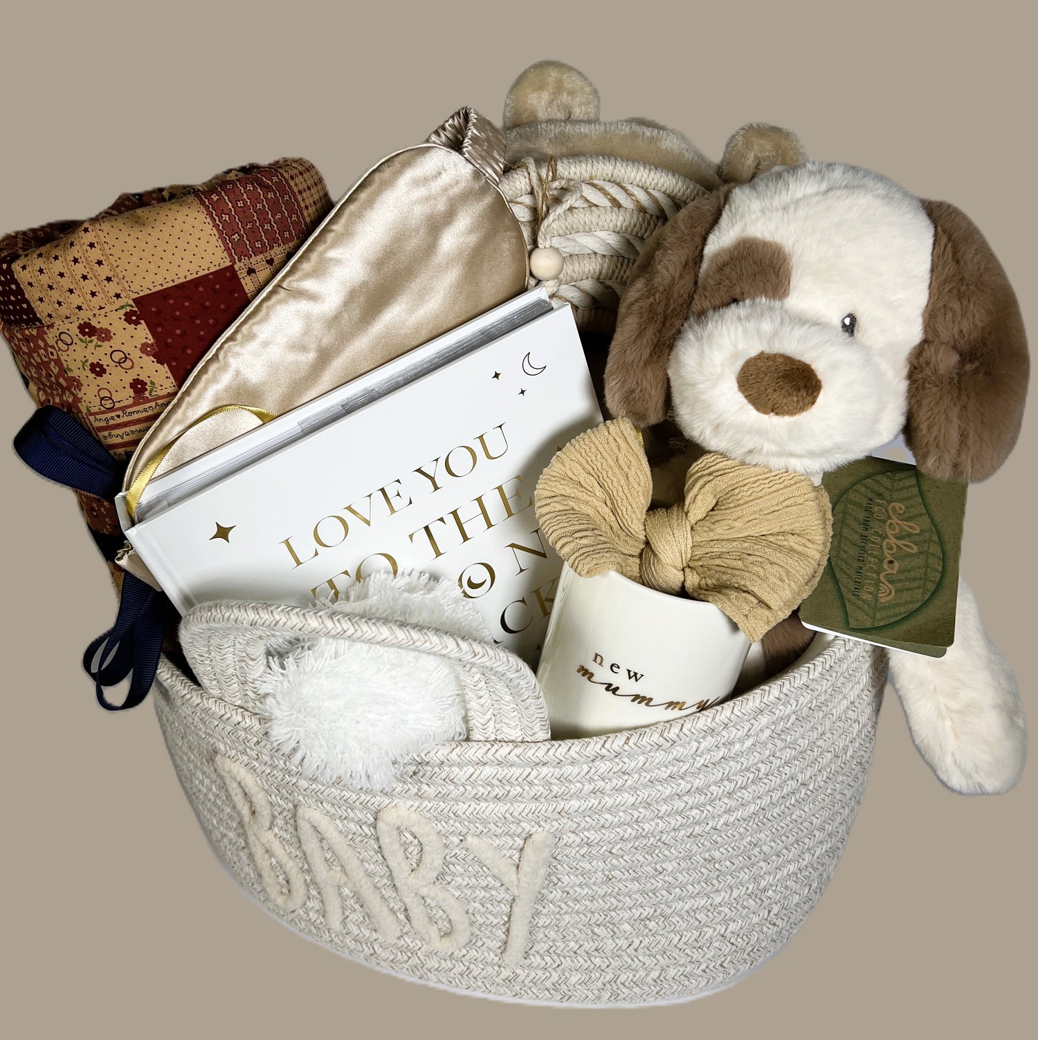 Nappy caddy baby hamper containing a tummy time blanket, baby photo album, ebbs puppy soft toy, new mummy mug, baby headband, baby dressing gown, baby pompom hat in white, a gold coloured silk adult eye mask, a nursery hanging decoration and some baby toiletries.