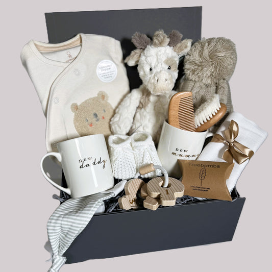 baby shower gift containing new mummy and daddy china mugs, a cotton baby sleepsuit, a baby brush and combs set, 1 baby pompom hat and a Mary Meyer Putty nursery giraffe soft baby toy