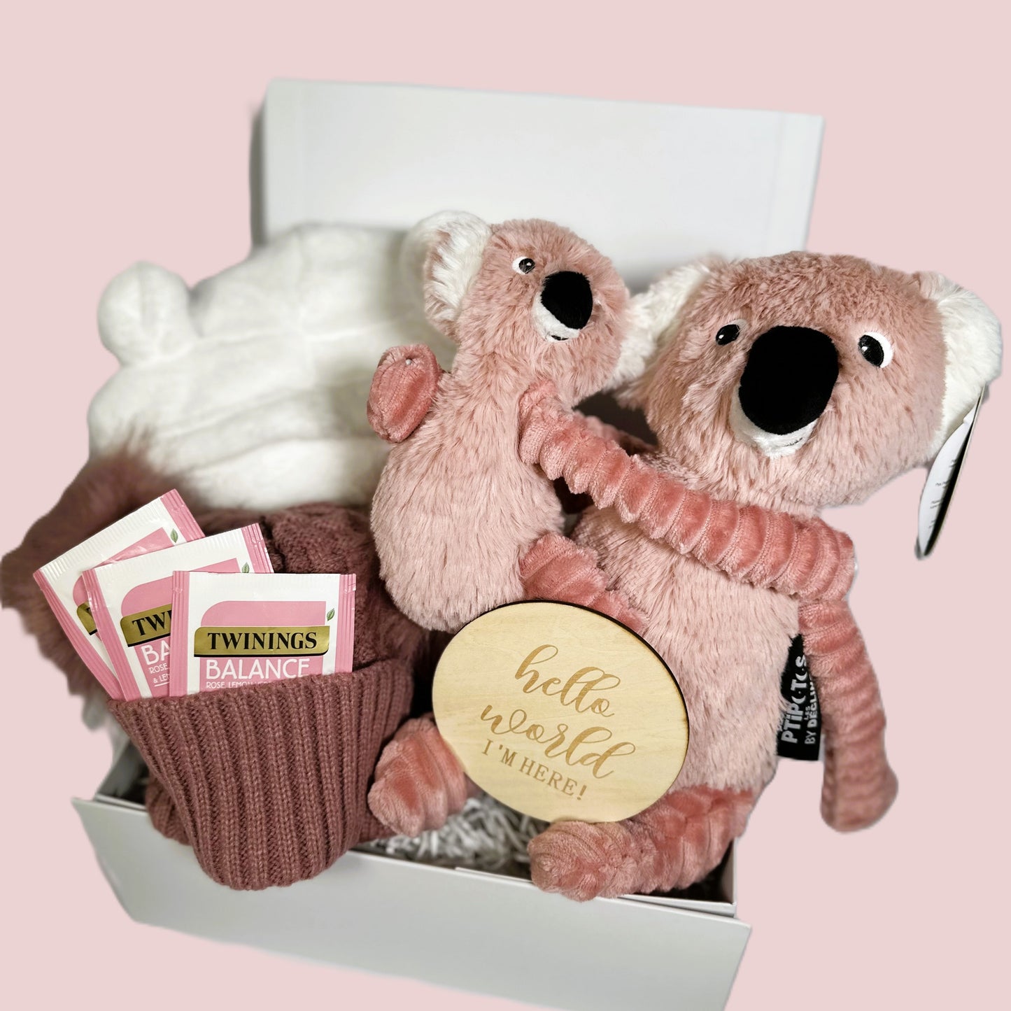 Pink baby girl hamper with a mummy Koala and her baby, a white hooded baby dressing gown, a dusky pink double pompom baby hat, Twinings teabags, a "Hello World" baby photograph sign and a white baby keepsake box.